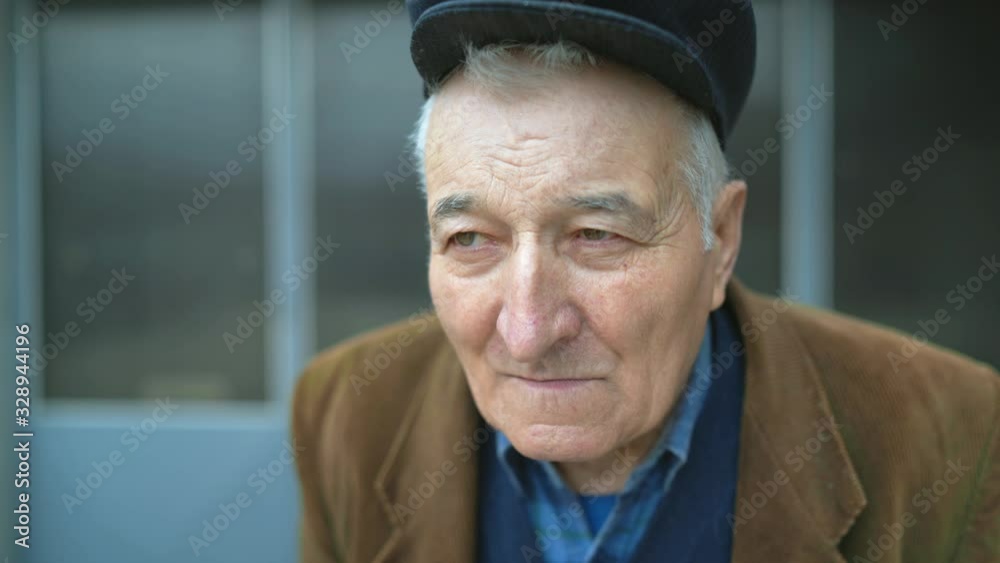 Portrait of senior man pensioner  looking around while standing alone outdoor wearing jacket and hat cap in front of house 