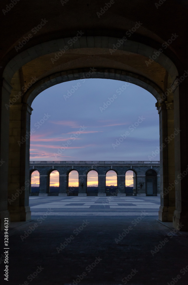 Dark tunnel leading to a large courtyard lined with arches in the middle of a colorful sunset