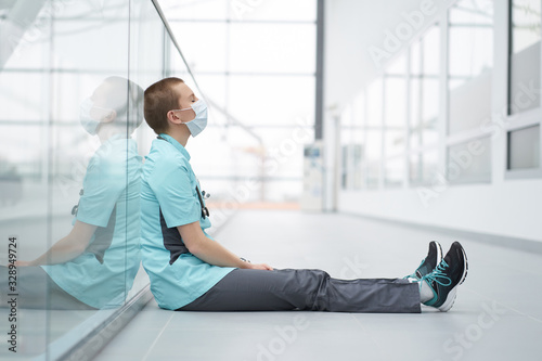 Burnout concept. Exhausted caucasian female short haircut doctor, wearing medical mask, sitting on a floor in corridor of clinic with closed eyes