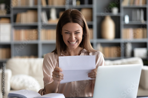 Excited young woman sitting at workplace at home, holding paper letter, received bank loan approval notice, job promotion. Happy businesswoman getting good news notification, money refund document.