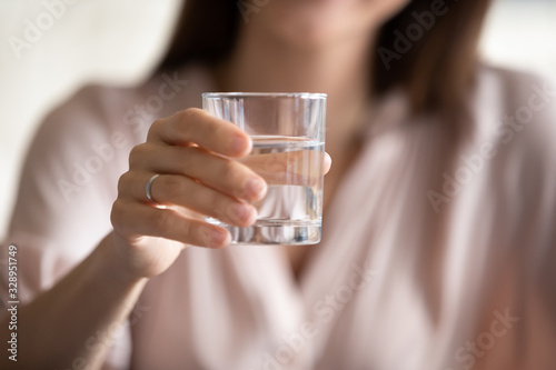 Cropped close up image focus on young woman reaching hand with glass of water to camera, proposing drinking fresh pure stilled liquid. Female nutritionist recommending daily rate, healthy habit.