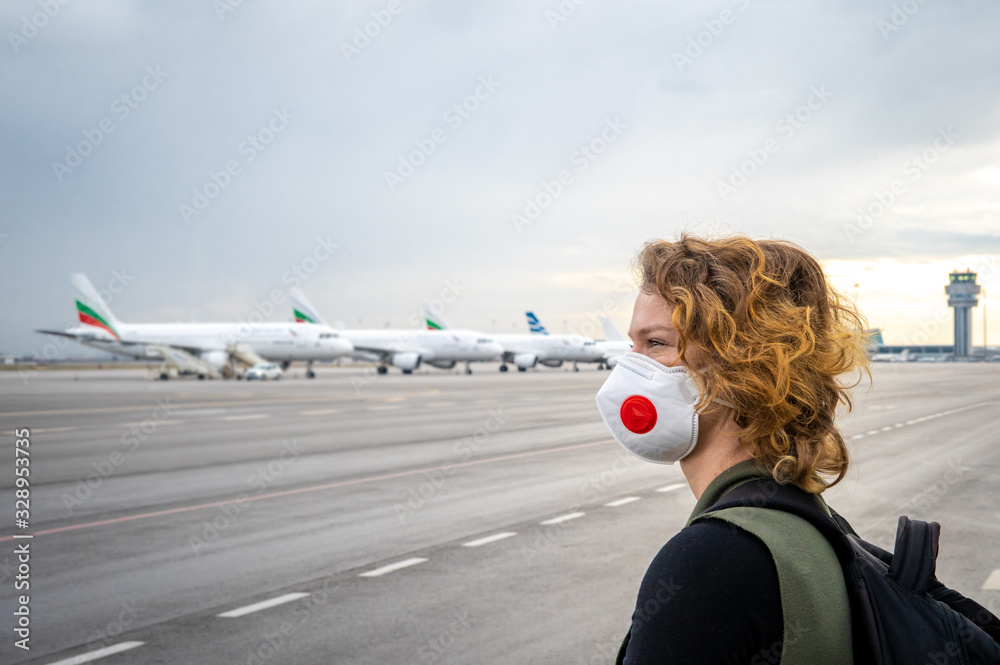 Woman with a mask on her mouth protects against the virus. Sadly looks at the aircraft parked at the airport. Canceled flights due to coronavirus - obrazy, fototapety, plakaty 
