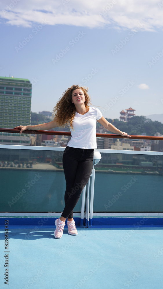 Happy beautiful girl on summer vacation on a cruise liner dreamily looks away and smiles against the backdrop of the port Asian city, a walk on the deck of the ship in sunny weather.