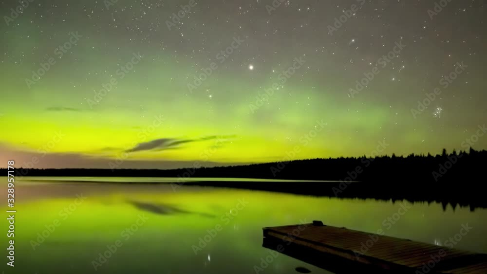 Time lapse of flashing vivid green Northern Lights reflecting in a calm lake with reeds moving slowly in the foreground and a line of pine and spruce trees along the horizon. Time lapse of flashing vivid green Northern Lights reflecting in a calm lake with reeds moving slowly in the foreground and a line of pine and spruce trees along the horizon.