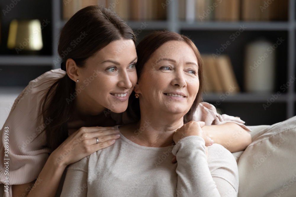 Fototapeta premium Head shot close up dreamy two generations women relaxing at home, looking aside. Young woman cuddling pleasant happy middle aged 60s mother, visualizing future, planning weekend leisure free time.