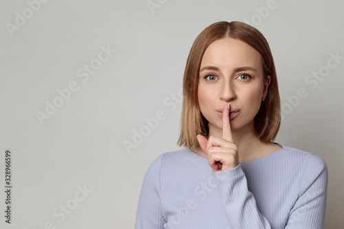 Shhh! European mysterious woman with short hair keep index finger over lips gesturing silence, isolated over grey background with copy space. Keep quiet, hush, privacy concept.  photo