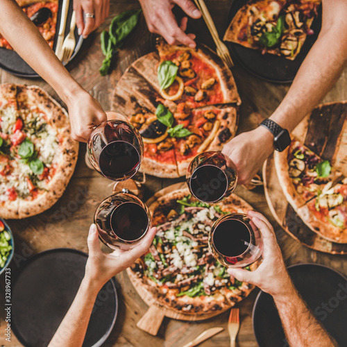 Family or friends having pizza party dinner. Flat-lay of people clinking glasses with red wine over rustic wooden table with various kinds of Italian pizza, top view, square crop. Fast food lunch