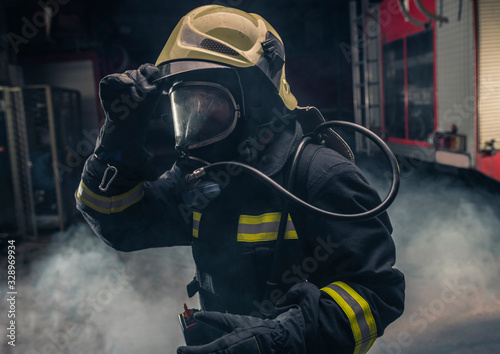 Portrait of a fireman wearing firefighter turnouts putting on oxygen mask. Dark background with smoke and blue light.