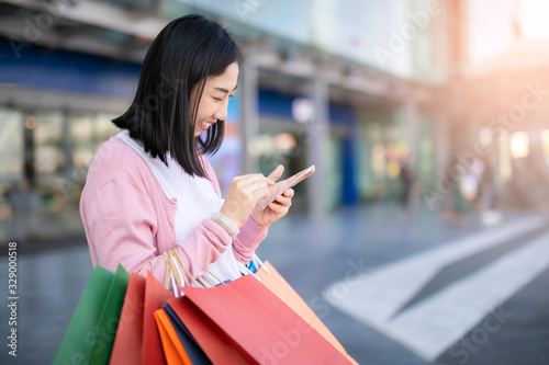 Happy Asian pretty girl holding shopping bags while using smartphone background shopping mall concept.