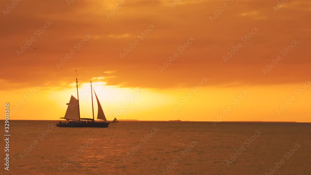 Sailing boats and ships are sailing through the calm ocean water at Key West in Florida, the southernmost point of the USA. The evening sky and clouds are colored orange and yellow toward the horizon.