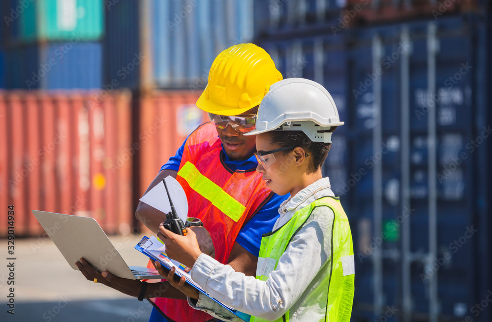 Worker man in hardhat and safety vest holding laptop and Female foreman talks on two-way radio control loading containers box from cargo
