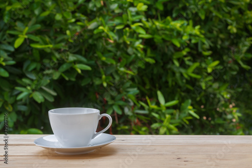 Image of clean empty white coffee cup or teacup and the plate put on old wooden table and blurry fresh green leaves tree in natural background with morning soft lighting and copy space.