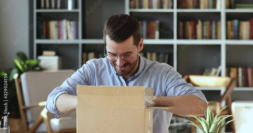 Smiling young man customer opening parcel cardboard box sitting at home office desk. Happy consumer unpacking postal shipping delivery satisfied with good purchase. Fast post shipment service concept