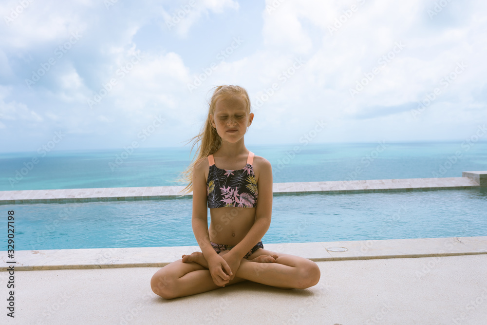 Little and beautiful girl sits by the pool in a bathing suit in lotus position. Morning yoga and calm. Sports practice and self-development. Healthy lifestyle Little and beautiful girl sits by the pool in a bathing suit in lotus position. Morning yoga and calm. Sports practice and self-development. Healthy lifestyle