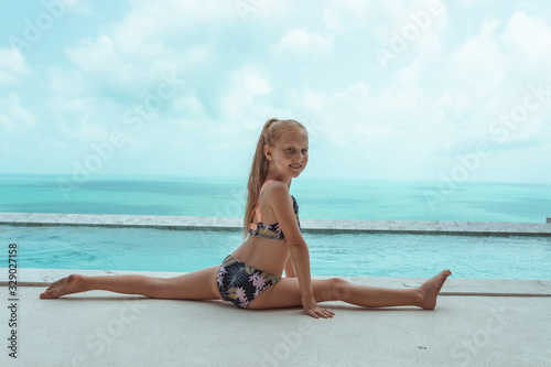 A small and beautiful girl with red hair and freckles sits by the pool in a bathing suit in the villa and poses in front of the camera. Children's fashion © Underwater girls