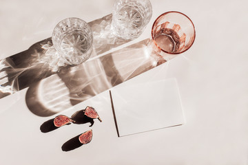Summer stationery still life scene. Glasses of water, cut figs fruit. Pink table background in sunlight. Blank business, greeting card, invitation mockup scene. Long harsh shadows. Flat lay, top view