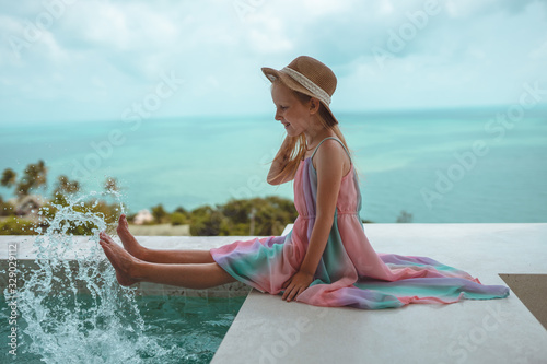 Little and beautiful girl with red hair and freckles sits by the pool in the villa and poses in front of the camera. Children's fashion
