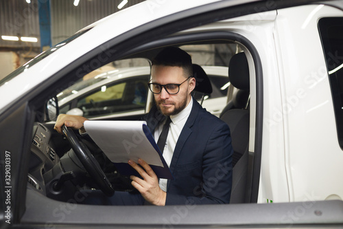 Young adult man wearing stylish suit and eyeglasses sitting in his car in auto service center reading cost sheet, horizontal medium portrait © AnnaStills