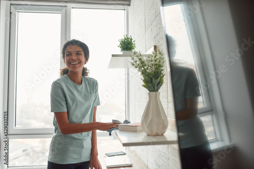 Making your home sparkling clean. Young happy afro american woman in uniform wiping dust off the shelves in the living room and smiling