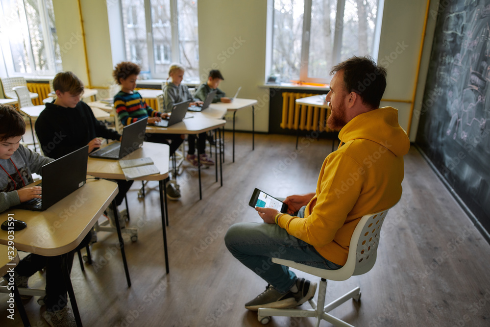 Where Learning Begins. Portrait of young male teacher communicating with his pupils, while sitting with tablet pc near the blackboard during a lesson in modern smart school
