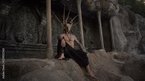 Tattooed masked skull ethnic pagan shaman sit on stage an ancient temple photo