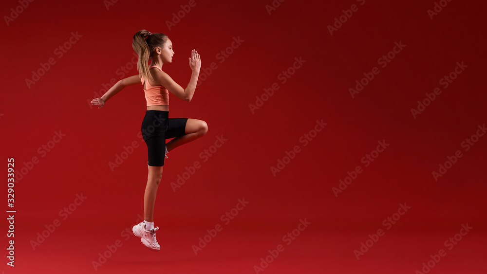 Higher and higher. A cute kid, girl is engaged in sport, she is looking away while jumping. Isolated on red background. Fitness, training, active lifestyle concept. Horizontal shot Higher and higher. A cute kid, girl is engaged in sport, she is looking away while jumping. Isolated on red background. Fitness, training, active lifestyle concept. Horizontal shot