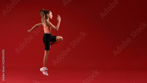 Higher and higher. A cute kid, girl is engaged in sport, she is looking away while jumping. Isolated on red background. Fitness, training, active lifestyle concept. Horizontal shot