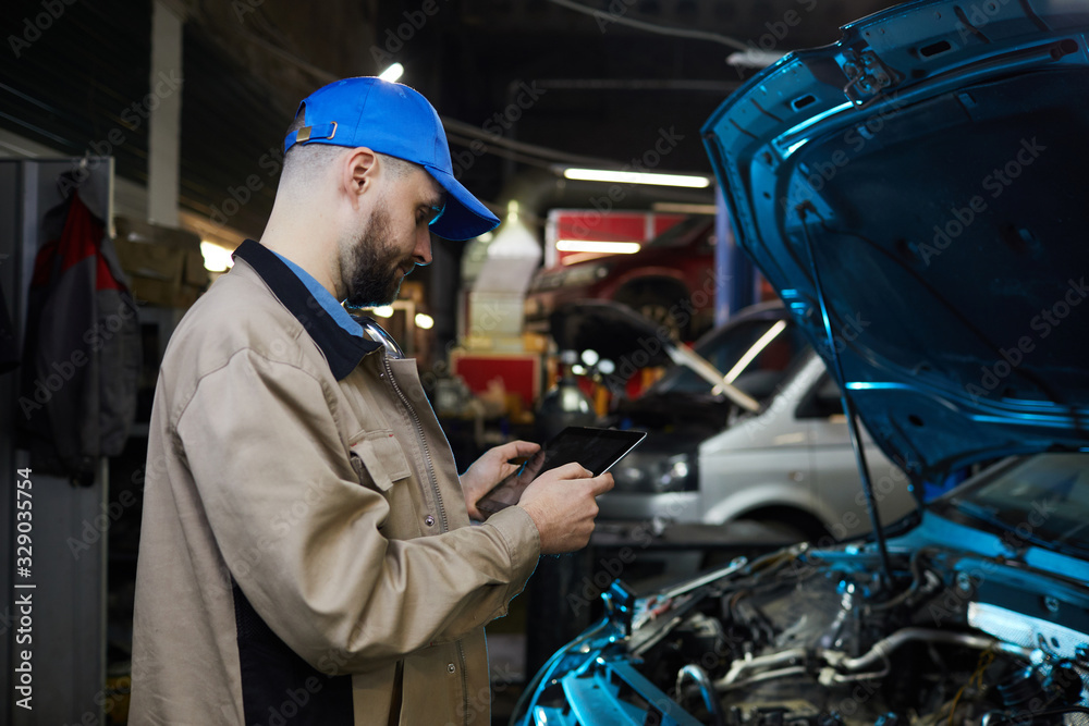 Cuacsian man with beard on face working in auto service workshop using tablet PC, horizontal side view shot Cuacsian man with beard on face working in auto service workshop using tablet PC, horizontal side view shot