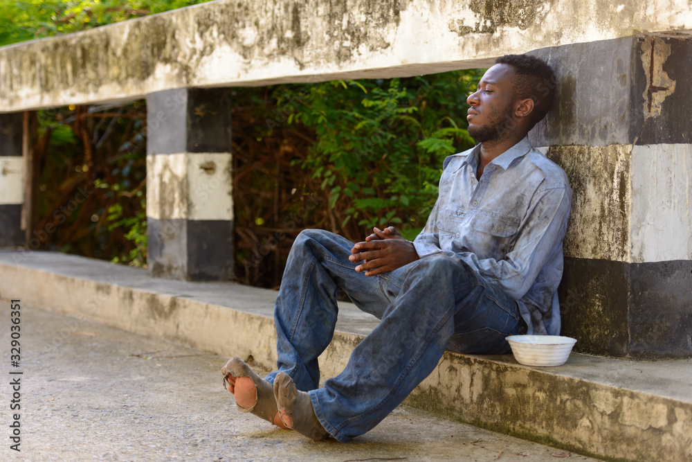 Young homeless African man with plastic bowl looking hungry