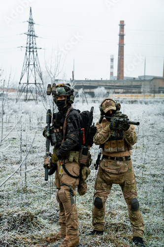 Men in camouflage cloth and black uniform with machineguns with factory on background
