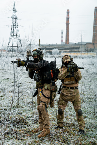 Men in camouflage cloth and black uniform with machineguns with factory on background. Soldiers with muchinegun aims aiming