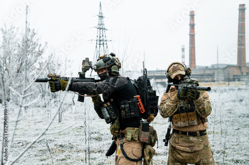 Men in camouflage cloth and black uniform with machineguns side to side with factory on background. Soldiers with muchinegun aims aiming