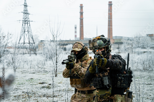 men in camouflage cloth and black uniform with machineguns side to side with factory on background. Soldiers with muchinegun aims aiming