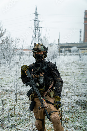 Man in black camouflage uniform hold machinegun. Soldier in the winter with factory on background