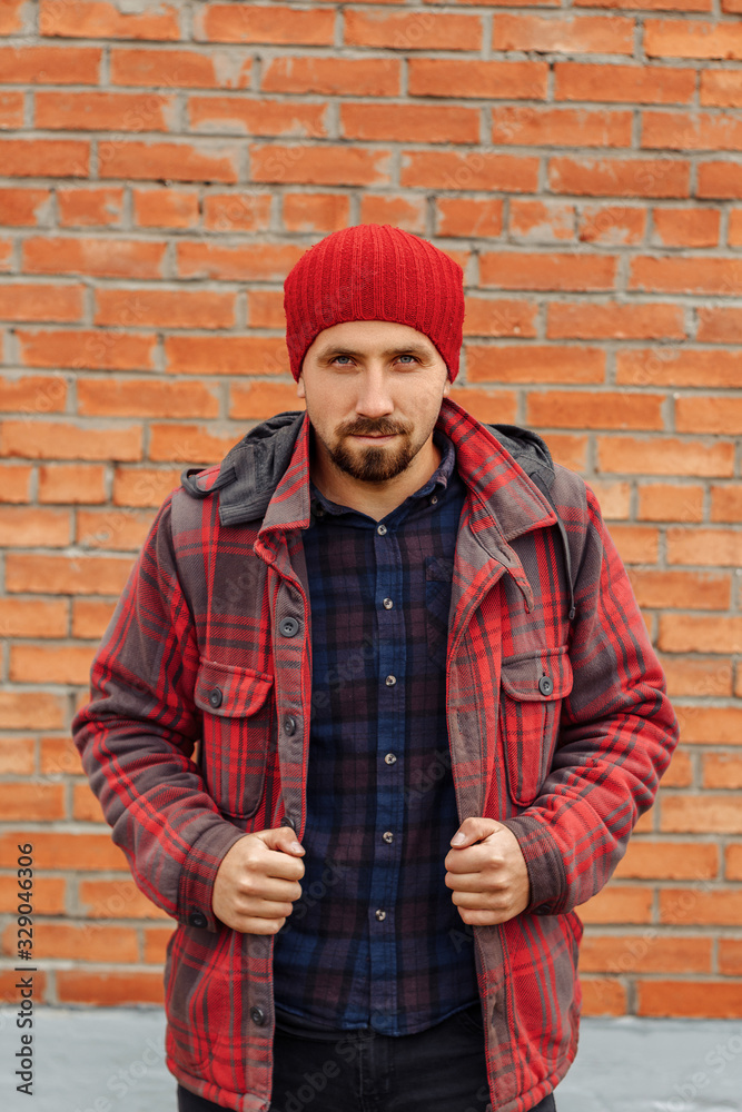 Young bearded man in red coat and hat. Guy with beard is standing in casual clothes against a brick wall and looking at camera. Young bearded man in red coat and hat. Guy with beard is standing in casual clothes against a brick wall and looking at camera.