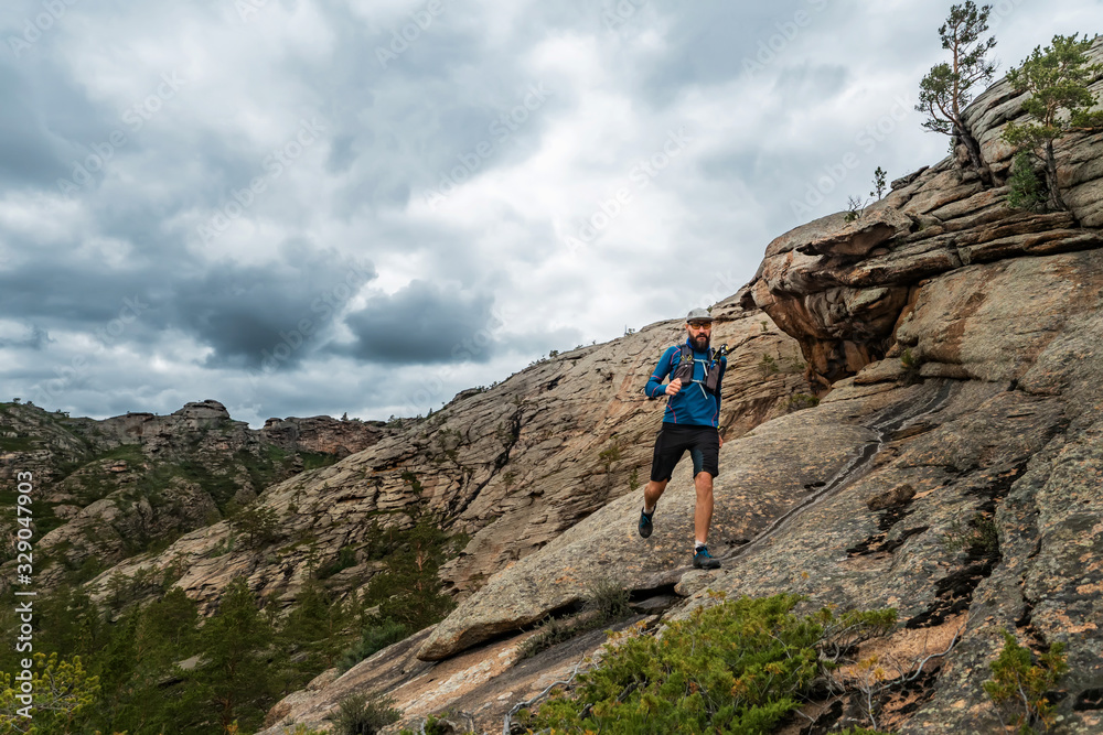Runner runs on the rocky mountains. A man in blue jersey and black shorts is training outdoors