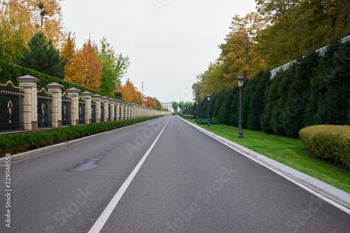 Grey smooth empty road. Decoratove fence, trees, bushes and pole lamps. photo