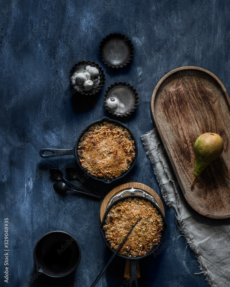 Two homemade crunchy pear tarts in black little pans surrounded by vintage molds filled with vanilla icecream and pear on a blue background