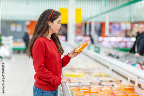 A young Caucasian woman in a red sweatshirt reads the information on the package with a semi-finished product. In the background is a supermarket. The concept of grocery shopping photo
