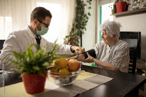Young serious Caucasian doctor wearing face mask adjusting blood pressure gauge on senior gray-haired woman's hand during house call medical check-up photo