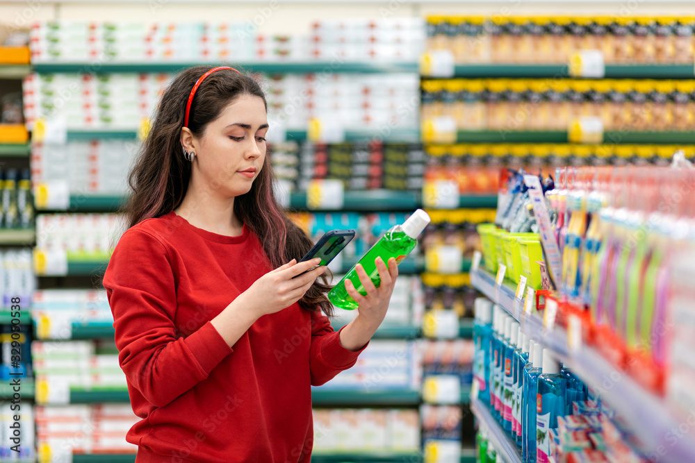 A young Caucasian woman scans the qr code on a shampoo bottle using her smartphone. In the background is a supermarket. Concept of modern technologies and shopping