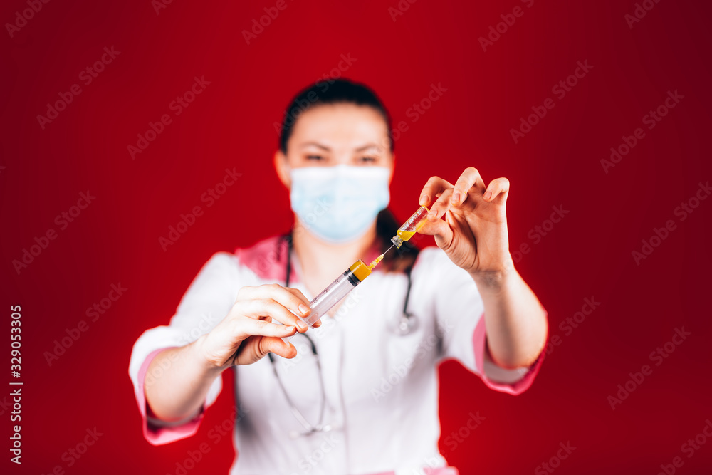 young girl doctor with a syringe in hand in a white coat and a mask on her face, fills a syringe on red background