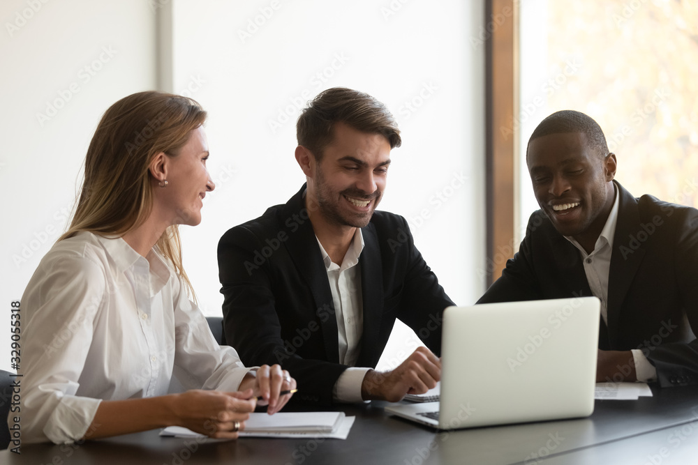 Happy mixed race business people sitting at table, looking at computer screen, satisfied with financial report, first project results. Smiling diverse colleagues having fun during break at office. Happy mixed race business people sitting at table, looking at computer screen, satisfied with financial report, first project results. Smiling diverse colleagues having fun during break at office.