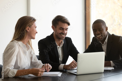Happy mixed race business people sitting at table, looking at computer screen, satisfied with financial report, first project results. Smiling diverse colleagues having fun during break at office.