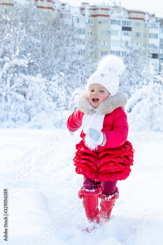 Enjoyment in the winter season. A beautiful little girl in a red coat and white hat with a scarf runs along a snowy path in a winter park. Smile and children's joy on the face. Great holidays. Snow