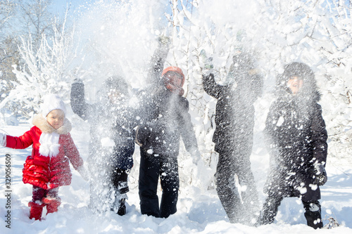 A group of children throw snow and enjoy the winter games in the fresh frosty air. Snow-covered trees behind them. Wonderful winter vacation. Healthy pastime for kids. Four boys and young little girl