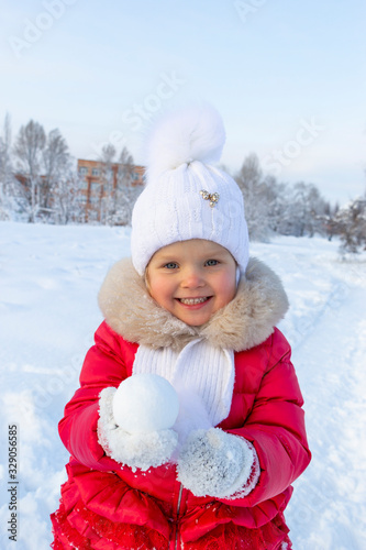 Little beautiful girl in a red coat and white scarf holds a round snowball in her hands and smiles. Snow-covered path and snowdrifts behind her. Children's entertainment during the winter holidays