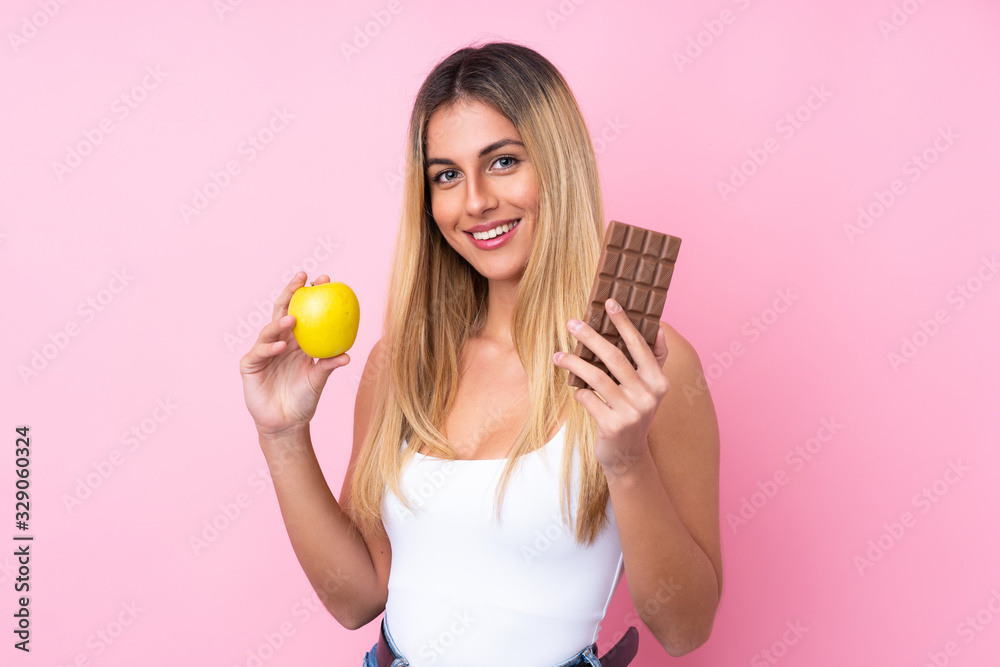 Young Uruguayan woman over isolated pink background taking a chocolate tablet in one hand and an apple in the other Young Uruguayan woman over isolated pink background taking a chocolate tablet in one hand and an apple in the other