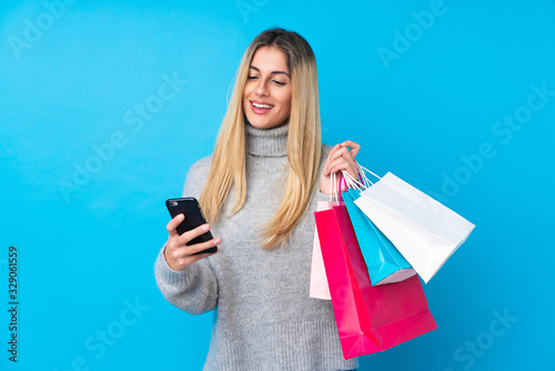 Young Uruguayan woman over isolated blue background holding shopping bags and writing a message with her cell phone to a friend