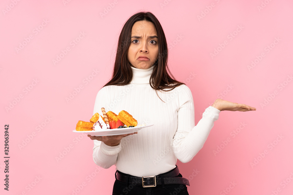 Young brunette woman holding waffles over isolated pink background unhappy for not understand something Young brunette woman holding waffles over isolated pink background unhappy for not understand something
